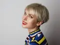Kobieta z blond włosami i grzywką baby bangs, w kolorowym golfie, uśmiecha się do aparatu.