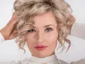 Blondynka z kręconymi włosami, z nonszalanckim gestem poprawiająca fryzurę. Jej oczy przyciągają uwagę, a usta są pomalowane na różowo.