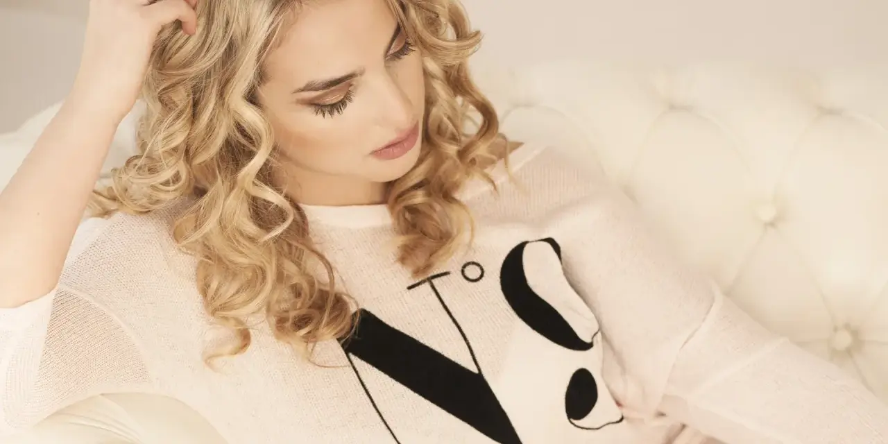 Kobieta z kręconymi włosami w stylu ombre blond, w jasnoróżowym swetrze z czarnym napisem, siedzi na białej kanapie.