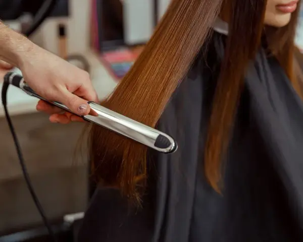 Stylista pokazuje, jak zrobić fale prostownicą, tworząc delikatne loki na długich, brązowych włosach.