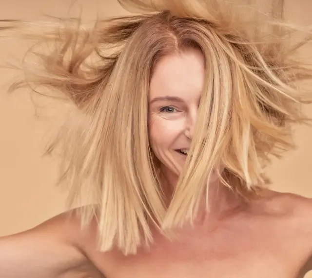 Kobieta z burzą włosów w odcieniu **kolor blond**, uśmiecha się, odsłaniając część twarzy.