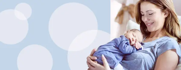 Uśmiechnięta mama tuli śpiącego noworodka w pasiastym pajacyku. Wskazówki, jak ubierać noworodka w domu.